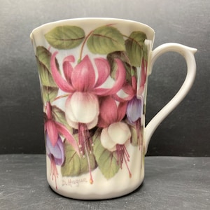 May include: White ceramic mug with a floral design featuring pink, white, and purple fuchsia flowers. The mug has a handle and the signature "D. Hague" is printed on the bottom.