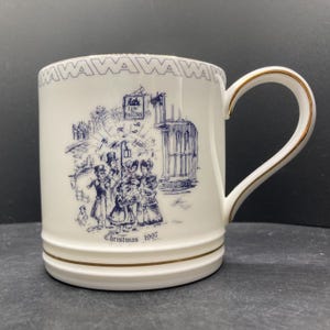 May include: White ceramic mug with a blue and gold design. The mug features a detailed blue illustration of a Christmas scene with figures and buildings. The handle and rim are trimmed with gold. The text "Christmas 1997" is printed on the mug.