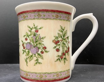 May include: A white ceramic mug with a floral design. The mug features a cream-colored background with illustrations of purple and red berries and green leaves. The top and bottom rims have a decorative border of red and purple.