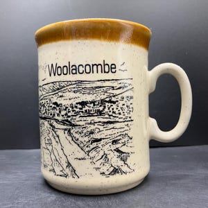 May include: Cream-colored ceramic mug with a brown rim and handle. The mug features a black and white illustration of a coastal town and the word "Woolacombe". The mug is speckled and has a curved handle.