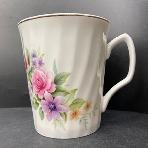 May include: White ceramic mug with a floral design featuring pink, purple, and yellow flowers. The mug has a gold rim and a handle.