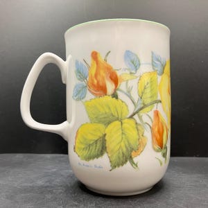 Może przedstawiać: Biały ceramiczny kubek z zakrzywionym uchwytem, ​​z kwiatowym wzorem żółtych i pomarańczowych pąków róż i zielonych liści. Obręcz ma jasnozielone wykończenie. Kubek ma gładkie, błyszczące wykończenie.