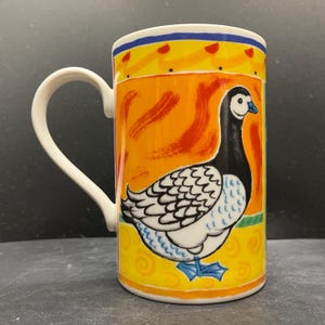 May include: A ceramic mug with a white handle and a colorful design. The mug features a goose illustration with black, white, and blue details against an orange background. The rim has yellow and blue accents.