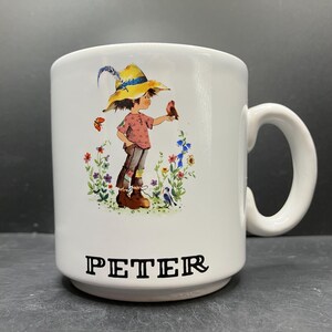 Op de afbeelding: Witte keramische mok met een grillige illustratie van een kind met een strohoed dat een vogel vasthoudt, omringd door bloemen en vlinders. De naam "PETER" is in zwarte blokletters op de bodem gedrukt. De mok heeft een gebogen handvat.