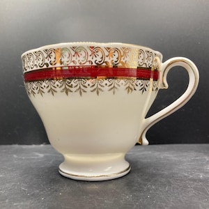 May include: Cream-colored teacup with a red band and gold accents. The cup features a decorative gold pattern around the rim and base, and on the handle. The handle and base are also trimmed with gold.