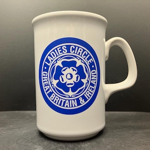 May include: White ceramic mug with a blue emblem reading "Ladies Circle Great Britain & Ireland." The emblem features a floral design. The mug has a curved handle and a cylindrical shape.