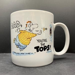 May include: White ceramic mug with a cartoon illustration of a man throwing darts. The mug has the text "DAD, YOU'RE THE TOPS!" in black lettering. The man is wearing a yellow and white striped shirt and blue pants.