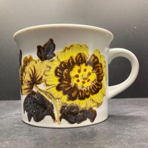 May include: White ceramic mug featuring a floral pattern in yellow, brown, and dark blue. The design includes detailed stylized flowers with a central pattern of red dots. The mug has a rounded handle.