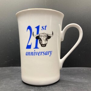 May include: White ceramic mug with a curved handle. The mug features blue text that reads "21st anniversary" with a bull graphic. The mug is a standard size for coffee or tea.