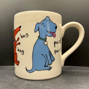 Könnte beinhalten: Cremefarbene Keramiktasse mit Cartoon-Hunde-Illustrationen. Ein Hund ist orange und bettelt, mit dem Wort "beg" zweimal geschrieben. Der andere Hund ist blau, keucht, mit dem Wort "pant" zweimal geschrieben. Die Tasse hat einen gebogenen Griff.