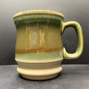 May include: A handmade ceramic mug with a rounded body and a sturdy handle. The mug features a layered design with shades of green, brown, and cream. The glaze gives a glossy finish to the stoneware. This mug is perfect for enjoying coffee or tea.