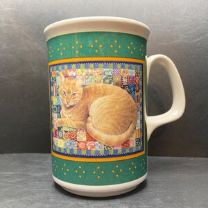 Op de afbeelding: Een witte keramische mok met een groen, geel en blauw patchwork ontwerp met een gemberkat. De kat ligt op een kleurrijk quilt.