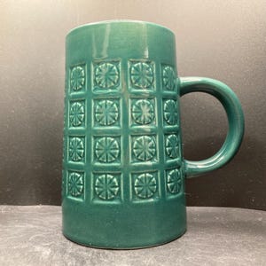 May include: A tall, emerald green ceramic mug with a large handle. The mug's surface is decorated with a repeating pattern of squares, each containing a stylized floral design. The mug is cylindrical and has a glossy finish.