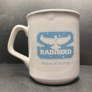 以下が含まれることがあります： 白いセラミック製のマグカップ。青いロゴには、スタイリッシュな鳥と「Rainbird」というテキスト、そして「Masters of Strategy.」というタグラインが書かれています。