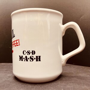 Peut inclure: Une tasse en céramique blanche avec une anse, ornée de texte en noir et rouge. On peut lire "COFFEE!" en rouge et "C*S*D M*A*S*H" en noir. La tasse présente un corps cylindrique à la finition lisse et une anse angulaire sur le côté droit.