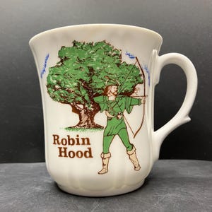 May include: White ceramic mug with a green and brown illustration of Robin Hood with a bow and arrow. The mug has the words "Robin Hood" printed in brown. The handle is white and curved.