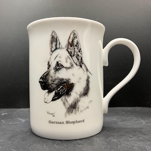 May include: White ceramic mug featuring a detailed black and white illustration of a German Shepherd dog. The mug has a curved handle and the words "German Shepherd" printed below the dog image.