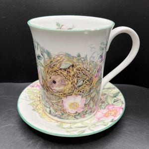 May include: White ceramic teacup and saucer set. The cup features a detailed illustration of a mouse in a nest, surrounded by flowers and foliage. The saucer mirrors the floral design with a green trim. A charming and delicate design.