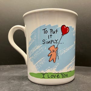 Könnte beinhalten: Weißer Keramikbecher mit gebogenem Henkel. Der Becher hat einen hellblauen Hintergrund mit einem Cartoon-Bären, der einen roten herzförmigen Ballon hält. Der Text auf dem Becher lautet "To Put It Simply... I Love You."
