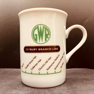 May include: A white ceramic mug featuring a green GWR circular logo above a brown horizontal bar labelled Tetbury Branch Line. Below the bar, a green line with tick marks lists station names: Tetbury, Trouble House Halt, Culkerton Halt, Church's Hill Halt, Rodmarton Platform, Jackaments Bridge Halt, and Kemble.
