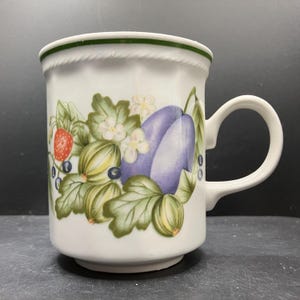 Könnte beinhalten: Weiße Keramiktasse mit Henkel, verziert mit einem farbenfrohen Obst- und Blumendesign. Das Design umfasst Erdbeeren, Blaubeeren, Stachelbeeren und lila Pflaumen mit grünen Blättern und weißen Blüten. Der Rand hat einen grünen Streifen.