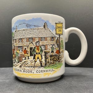 Op de afbeelding: Witte keramische mok met een illustratie van de Jamaica Inn in Bodmin Moor, Cornwall. Het ontwerp toont een man in een groene jas en gele broek, en de tekst "Jamaica Inn" op een bord.