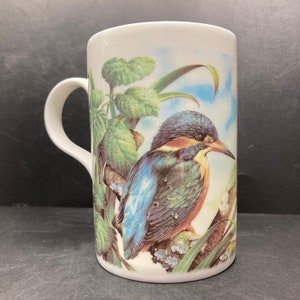 May include: A white ceramic mug with a blue, green, and orange kingfisher bird perched on a branch with green leaves. The background is a light blue sky.
