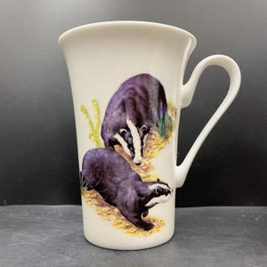 May include: White ceramic mug featuring two badgers in a naturalistic setting. The badgers are depicted with black and white markings and are set against a backdrop of yellow and green foliage. The mug has a curved handle.