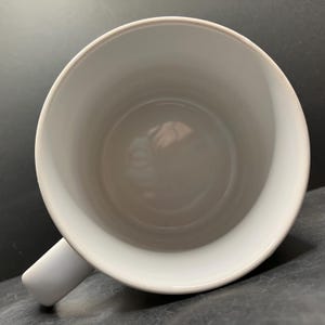 Puede incluir: Una taza de cer&aacute;mica blanca con un asa curva. El interior de la taza es blanco y liso. La taza est&aacute; vac&iacute;a y parece limpia. La taza se ve desde arriba, mostrando el interior y el asa.