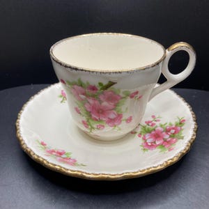 May include: A cream-coloured teacup and saucer set, decorated with pink floral motifs and gold edging. The teacup features a curved handle, and the saucer is round. The set is likely made of porcelain.
