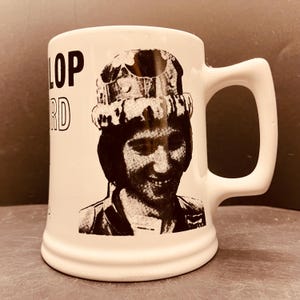 May include: A white ceramic mug featuring a black graphic print of a person wearing a crown. The text "LOP" and "RD" is printed in black on the left side of the mug. The mug has a smooth, cylindrical body with a sturdy, angular handle on the right side.
