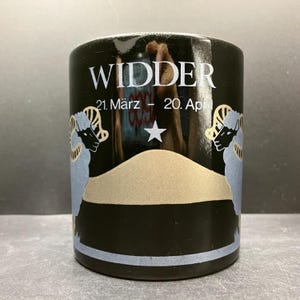 以下が含まれることがあります： 白い「WIDDER」の文字と「21. März - 20. April」の日付が入った黒いセラミックマグ。金と青の星と牡羊座のイラストが特徴です。マグカップの底には水色のストライプが入っています。