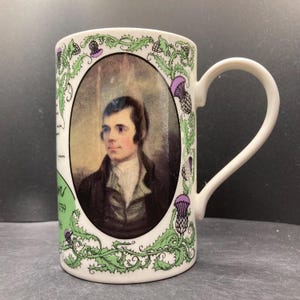 以下が含まれることがあります： 楕円形のフレームに男性の肖像画が描かれた白いセラミックマグ。マグカップは、緑と紫のアザミのデザインと緑のボーダーで装飾されています。マグカップには白いハンドルが付いています。