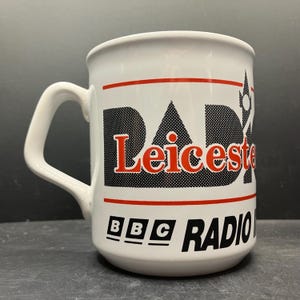 May include: White ceramic mug with a curved handle. The mug features the text "DAD Leicester" in a black and red design, with "BBC RADIO" below. The mug has a glossy finish.