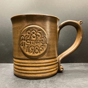 May include: Brown ceramic mug with a handle. The mug features the embossed text "985 Harwell 1985" within a circular design. The mug has horizontal ridges near the base and a small decorative element at the handle's base.