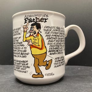 May include: White ceramic mug with a handle, featuring a cartoon illustration of a man in a yellow shirt and brown trousers. The mug has the text "Anatomy of a Father" and humorous text about fathers.