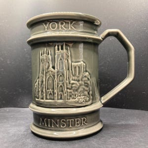 May include: Gray ceramic mug with a handle, featuring a detailed relief of a cathedral. The mug is inscribed with "YORK" at the top and "MINSTER" at the base. The design includes architectural details and a small car.