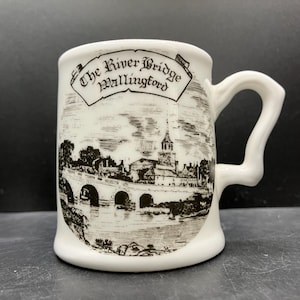 May include: White ceramic mug with a curved handle. The mug features a black and white illustration of a bridge and buildings, with the text "The River Bridge Wallingford" in a banner across the top.
