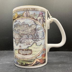 May include: White ceramic mug featuring a vintage world map design with the text "HYDROGRAPHICA TABULA". The mug has a curved handle and a tag attached. The map includes illustrations of people and animals.