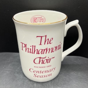 May include: White ceramic mug with a gold rim, featuring the text "The Philharmonic Choir" in burgundy script. The mug also includes the text "Founded 1896" and "Centenary Season". A musical note logo is at the top.