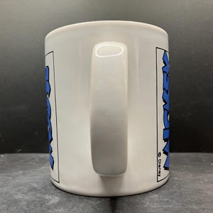 Puede incluir: Taza de cer&aacute;mica blanca con un asa grande. La taza presenta la palabra "Mickey" en azul, delineada en negro, en ambos lados. El logotipo de Disney es visible en el lado derecho de la taza.