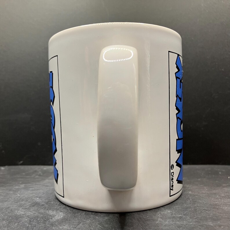 Puede incluir: Taza de cer&aacute;mica blanca con un asa grande. La taza presenta la palabra "Mickey" en azul, delineada en negro, en ambos lados. El logotipo de Disney es visible en el lado derecho de la taza.