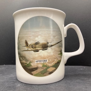 May include: White ceramic mug with a detailed image of a Spitfire aircraft flying above a landscape. The mug has a curved handle and the word "SPITFIRE" is printed below the plane.