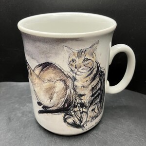 May include: White ceramic mug featuring a detailed sketch of a cat in shades of brown, gray, and black. The cat is depicted in a relaxed pose, with its body and tail visible. The mug has a curved handle and a smooth, glossy finish.