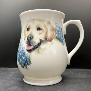 May include: White ceramic mug featuring a smiling yellow Labrador Retriever. The mug has a curved handle and is decorated with blue floral accents. The dog's portrait is detailed, with a pink tongue visible.