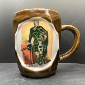 Op de afbeelding: Een bruine keramische mok met een centrale illustratie van een persoon in traditionele Schotse kleding, inclusief een groene tartan kilt en plaid, met de tekst "CAMPBELL OF BREADALBANE" in het zwart onderaan de afbeelding.