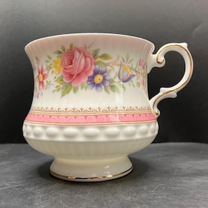 May include: A white porcelain teacup featuring a floral pattern with pink roses, blue and yellow flowers, and green leaves. The cup has a scalloped rim, a pink band with gold trim, a textured beaded middle section, and a gold-trimmed ornate handle.