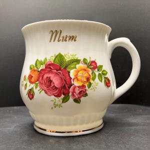 May include: Cream ceramic mug with a floral design of red, orange, and yellow roses, green leaves, and the word "Mum" in gold script. The mug has a gold rim and a curved handle.