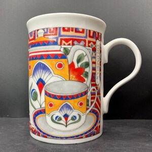 May include: White ceramic mug with a colorful design featuring a teapot, cup, and saucer. The design includes red, blue, yellow, and green patterns. The word "COLOMBIAN" is printed vertically on the side of the mug.