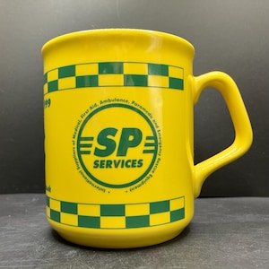 May include: A yellow ceramic mug with a green and white checkered pattern around the top and bottom. The mug features the text "SP SERVICES" in a circular logo, with additional text indicating it is for medical, first aid, ambulance, paramedic, and emergency rescue equipment.